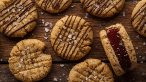 A flat lay of creative peanut butter cookie variations — drizzled with chocolate, sprinkled with sea salt, sandwich-style with jam filling, all on a rustic wooden surface with cozy kitchen lighting.