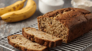A freshly baked banana bread loaf cooling on a wire rack, sliced open to show moist texture, with bananas and flour in the background.