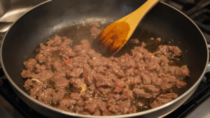 A frying pan on a stovetop, sizzling ground beef being stirred with a wooden spoon.