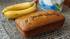 A loaf of golden banana bread cooling on a kitchen counter, with ripe bananas and a bag of flour beside it.