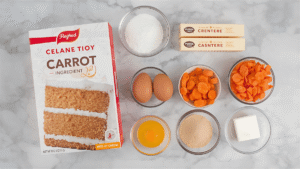 A top-down shot of all carrot cake ingredients on a marble countertop — cake mix box, eggs, grated carrots, butter, cream cheese, and sugar.