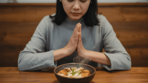 A woman sitting at a wooden dining table with a bowl of steaming food, hands clasped around it, eyes closed in gratitude before taking a bite.