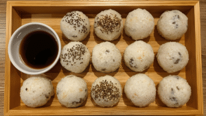 A wooden tray of freshly made rice balls — some plain, some wrapped in nori, sprinkled with sesame seeds, next to a small bowl of soy sauce.