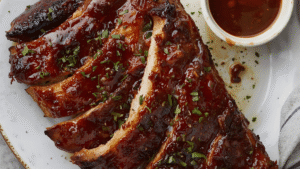 Finished ribs on a serving plate, glistening with caramelized brown sugar glaze, sprinkled with fresh herbs, next to a small bowl of barbecue sauce.