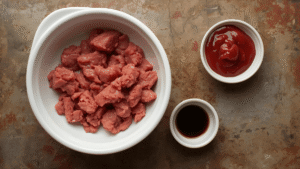 Ingredients flat lay — ground beef in a bowl, ketchup in a small dish, Worcestershire sauce in a tiny cup, all on a rustic kitchen counter.