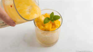 Pouring creamy mango mixture into a glass over cooked sago pearls, topped with mango cubes and a mint sprig, white marble table background.