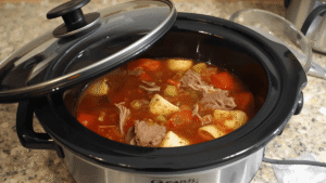 Slow cooker on a kitchen counter, lid lifted to reveal hearty soup bubbling inside with brisket and vegetables.