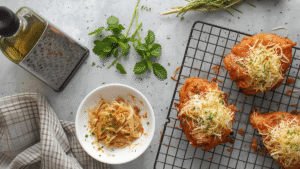 Top-down image of kitchen tools: grater, olive oil bottle, herbs, and a cooling rack with cooked chicken parmesan resting before serving.