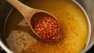 Wooden spoon stirring red pepper flakes into golden honey in a saucepan.