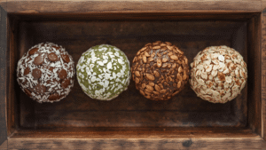 A flat lay of assorted energy ball variations — chocolate, coconut, matcha, and oat — arranged in a rustic wooden tray with natural lighting and shallow depth of field, realistic photography.