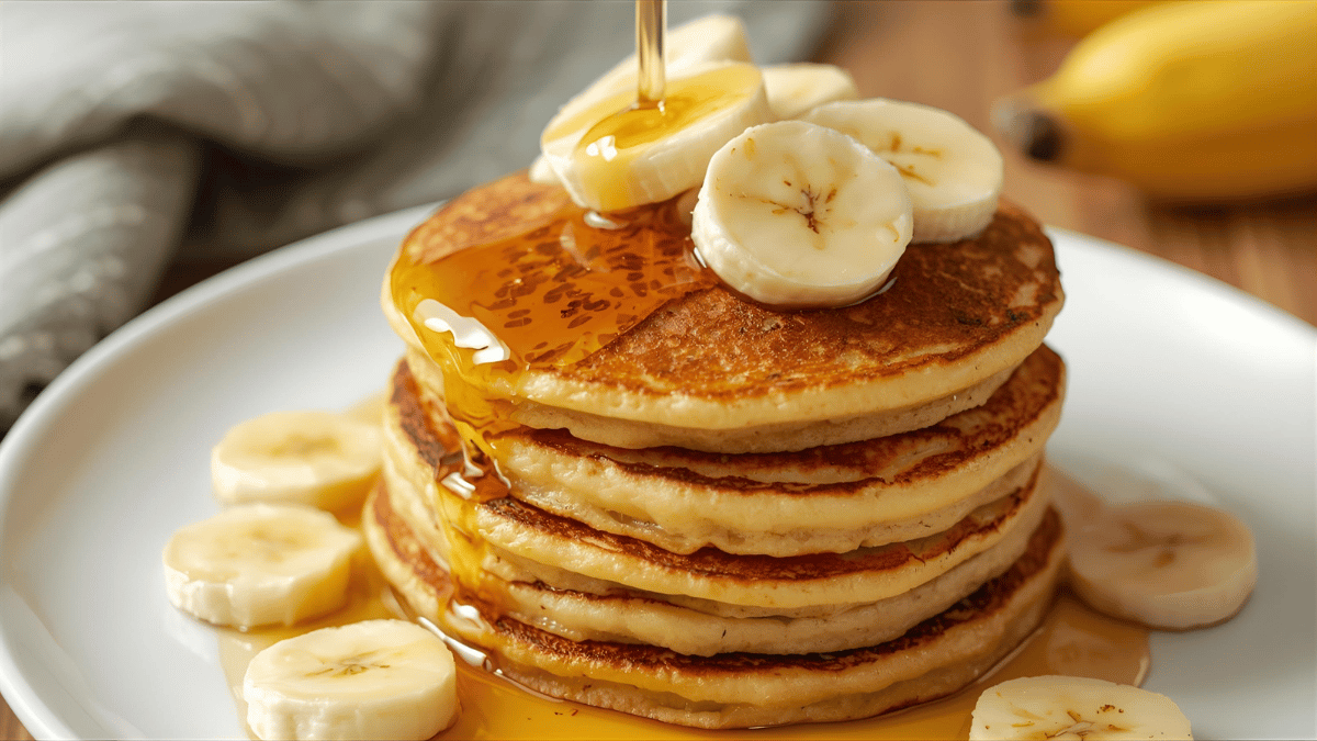 A stack of golden banana pancakes topped with honey and slices of banana.
