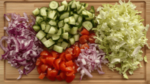 Chopped fresh vegetables on a wooden board: cucumber, cherry tomatoes, red onion, and shredded lettuce ready for a beef pita wrap.