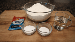 Close-up shot of bread ingredients: flour, instant yeast packet, warm water in a glass, sugar, salt, and oil on a wooden kitchen counter.
