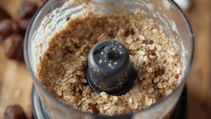 Macro shot of blended mixture for energy balls in a food processor bowl, showing creamy texture with oats and date bits visible, natural kitchen background, daylight tone, realistic photography.