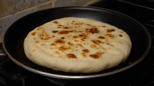 Soft pita bread warming on a skillet, slightly puffed and golden, rustic kitchen background.